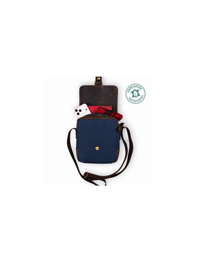 A navy blue canvas crossbody bag with dark brown leather trim and a gold snap button, shown open with its strap laid out. A small white dice-rolling prop is tucked inside, and a green circular certification badge is displayed to the right on a plain white background.