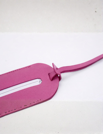 A close-up of a bright pink leather luggage tag with a zippered slot and a matching leather loop strap, set against a plain white background.