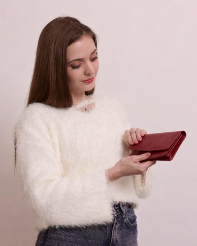 A woman with long brown hair wearing a fluffy white sweater and blue jeans, holding a dark red leather clutch wallet against a plain light background.