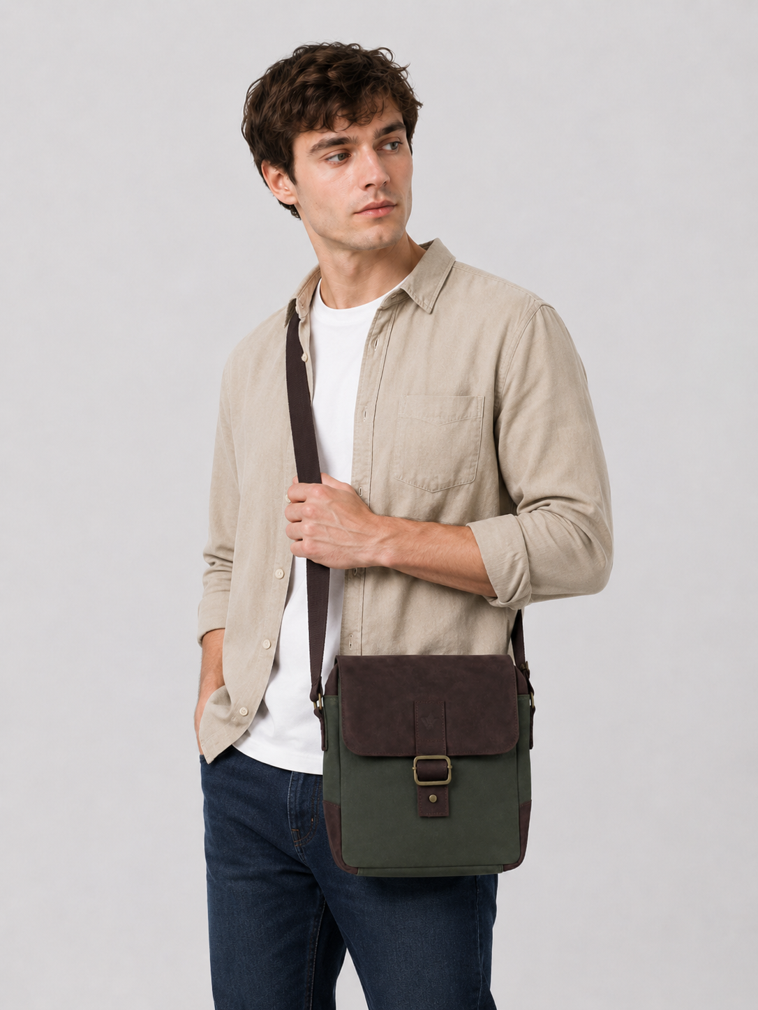 A man wearing a beige button-up shirt, white t-shirt, and dark blue jeans models a green and brown crossbody messenger bag against a light gray studio background.
