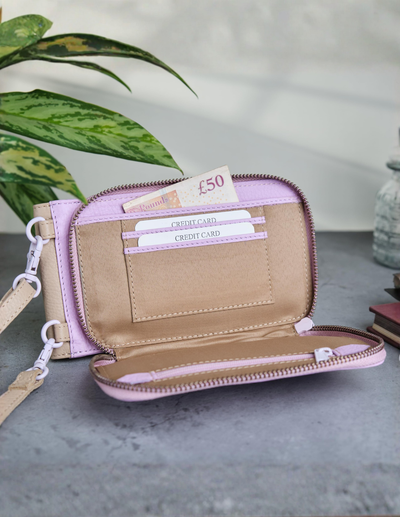 A light pink zip-around wallet shown open, revealing a tan interior with card slots, a £50 note, and a wristlet strap. The wallet is styled on a gray surface with a green plant leaf and a small vase in the background.