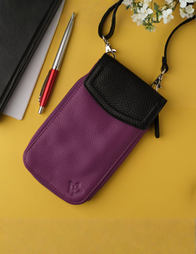 A purple and black pebbled leather phone pouch with a black strap, styled on a yellow background with a pen, notebook, and small white flowers.