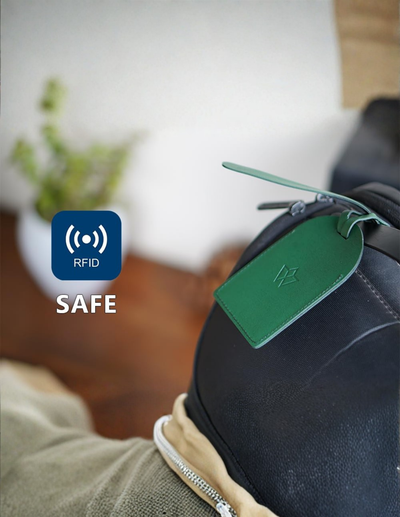 A close-up of a dark backpack with a green RFID luggage tag attached, with a blue 'RFID SAFE' graphic overlay on the left. The background is a softly blurred indoor setting with a wooden floor and a plant.