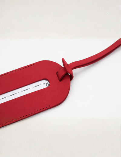 A close-up of a red leather luggage tag with a white insert slot and a matching red strap, photographed on a clean white background.