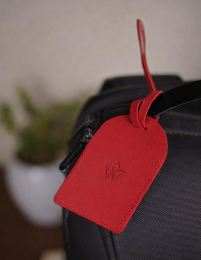 A bright red leather luggage tag with an embossed logo, attached to a black backpack strap, with a softly blurred indoor background.
