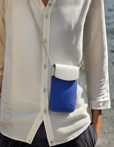 A close-up of a person wearing a white textured button-up shirt and dark pants, carrying a small crossbody pouch bag with a white flap and a vibrant blue body.