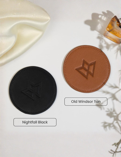 Two circular leather-like coaster or mat samples on a white background: one black labeled 'Nightfall Black' and one tan labeled 'Old Windsor Tan,' with decorative fabric and a small bottle in the corners.