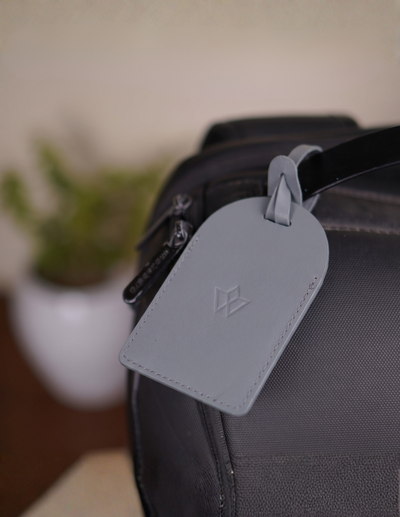 A light gray leather luggage tag with an embossed geometric logo, attached to a black textured bag strap, shown in a close-up shot with a blurred indoor background.
