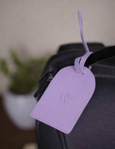 A lavender purple leather luggage tag with an embossed LV logo, hanging from a black bag strap in a softly blurred indoor setting.