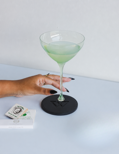 A hand with black fingernails holds a clear stemmed coupe glass containing a pale green drink, resting on a round black coaster. A small branded card is visible on the table to the left against a clean white background.
