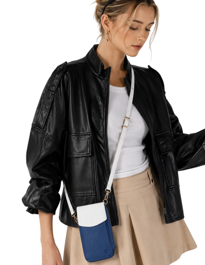 A woman wearing a black faux-leather jacket, white top, and beige pleated skirt, carrying a blue and white color-blocked crossbody phone bag with a white strap.