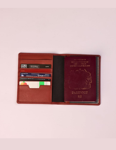 A brown leather passport holder shown open, containing card slots with sample cards, a black interior lining, and a burgundy passport insert, photographed on a light pink background.