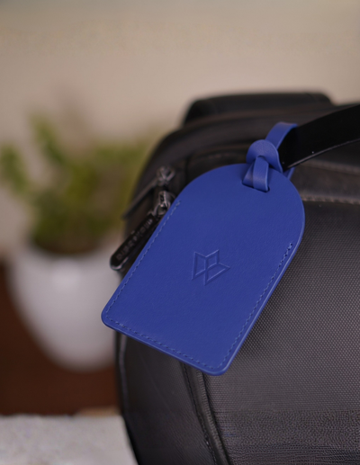 A bright blue leather luggage tag with an embossed geometric logo, attached to a black textured bag strap in a softly blurred indoor setting.