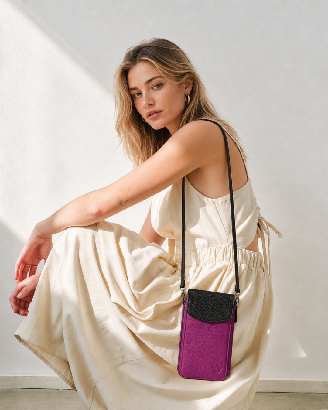 A woman in a cream-colored dress models a purple and black crossbody phone bag against a white wall.