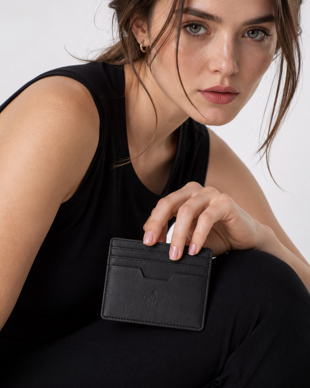 A close-up of a woman in a black sleeveless top holding a slim black leather card holder wallet against a light gray background.