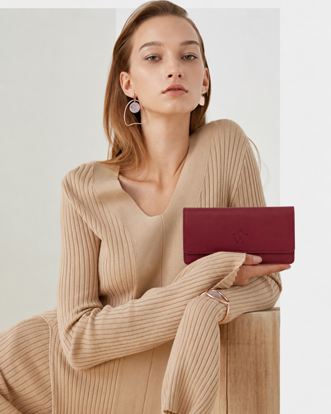 A woman wearing a beige ribbed knit sweater holds a burgundy leather clutch bag with a small star logo, posed against a light gray background.