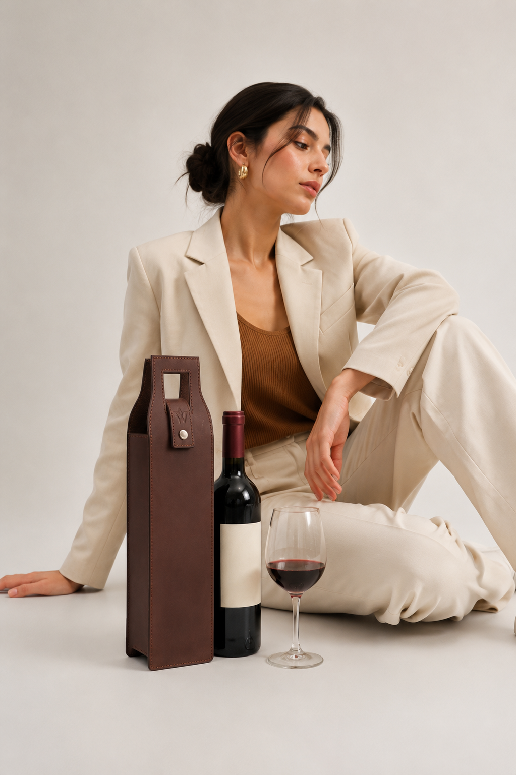A woman in a beige blazer and matching pants sits on a light studio floor beside a brown leather wine tote, a dark wine bottle, and a glass of red wine.