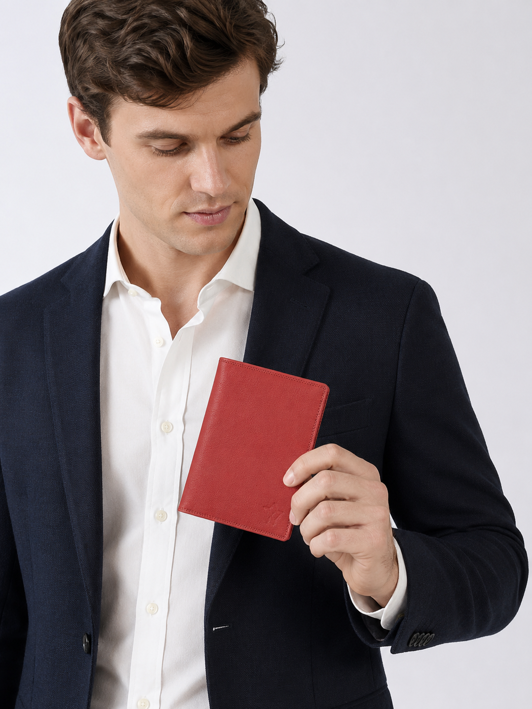 A man in a navy blue suit jacket and white dress shirt holds a small red leather notebook or passport cover against a light gray studio background.
