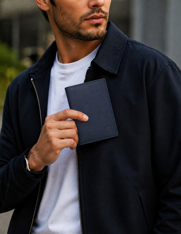 A man wearing a dark navy blue zip-up jacket over a white t-shirt, holding a matching dark navy leather passport cover or notebook in his hand.