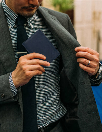 A man wearing a dark gray suit jacket, black tie, and blue-and-white check shirt holds open his jacket to reveal a navy blue leather passport cover or wallet.
