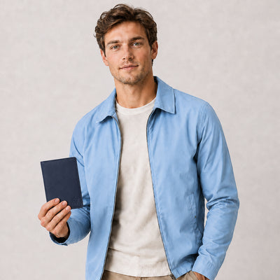 A man wearing a light blue overshirt over a cream knit sweater and khaki pants, holding a dark blue passport against a light gray textured background.
