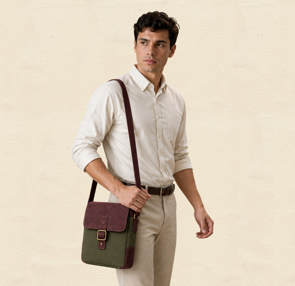 A man wearing a cream dress shirt and beige trousers carries a green and burgundy crossbody bag with a brown leather strap against a textured beige background.
