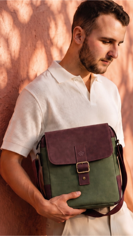 A man in a white polo shirt stands against a textured terracotta wall with leaf shadows, holding a green canvas messenger bag with a dark purple leather flap.