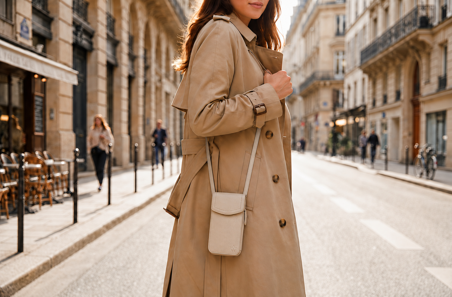 A woman wearing a classic beige trench coat and carrying a small beige shoulder bag, standing on a city street with buildings in the background.